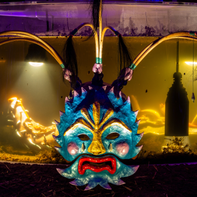 A brightly coloured theatrical mask with blue, gold and red features is displayed in front of a large illuminated tank filled with amber-coloured water. Curved poles and strands of dark hair extend from the top of the mask, creating a dramatic, horn-like shape. Warm yellow lighting inside the tank contrasts with the dark surroundings, highlighting reflections, suspended objects and organic materials within the display. The scene has an immersive, artistic atmosphere with vivid colours and low-light exhibition lighting.