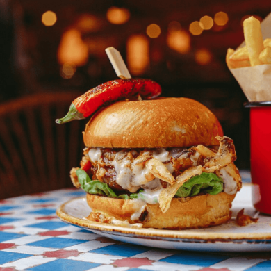 A large burger on a plate with cheese and salad on it. There is a red chilli on top of the burger. On the plate next to the burger is a red pot of chips. The table the plate is on is decorated with red and blue squares.