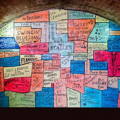 Colourful mural displayed within a brick-arched alcove, featuring hand-painted names of famous music artists and bands associated with Liverpool and The Cavern Club. The artwork is made up of brightly coloured blocks in shades of blue, pink, orange, green and yellow, with names including The Beatles, Gerry and the Pacemakers, Queen, Oasis, The Rolling Stones, The Who, Arctic Monkeys and Cilla Black. Warm lighting highlights the painted wall against the dark red-brown brick surroundings.