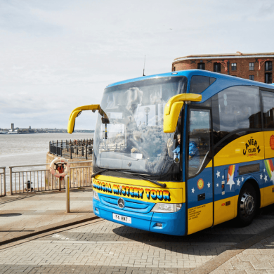 A blue and yellow bus with 'Magical Mystery Tour' on the front and side. It is infront of a river and an old brick warehouse building.