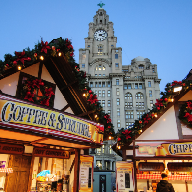 Festive market stalls decorated with garlands, lights and red poinsettias stand in front of Liverpool’s Royal Liver Building at dusk. Signs advertising coffee, strudel and Christmas bakery treats glow warmly, creating a seasonal atmosphere in the city centre.