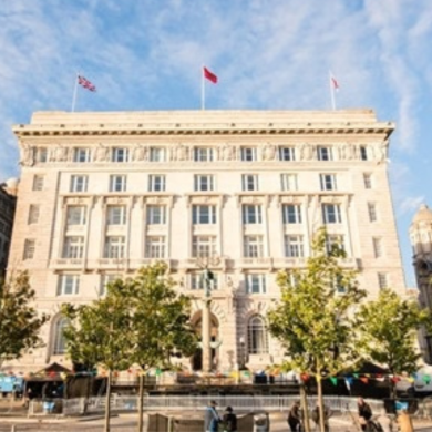 Cunard Building, the former headquarters of White Star Line in Liverpool, viewed from the waterfront. The historic stone building stands among the city’s Three Graces, with trees and public space in the foreground beneath a bright blue sky.