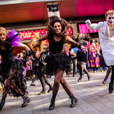 A group of performers in elaborate Halloween costumes dance in a public plaza outside Liverpool ONE. Dressed in black gothic outfits and zombie-style makeup, the performers leap and move energetically while crowds gather behind them to watch. Bright pink shopfront lighting and dramatic movement create a lively festival atmosphere.