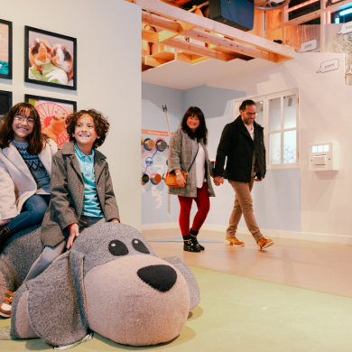 Two children sit on a large soft sculpture shaped like a grey dog inside an interactive family exhibition space. Behind them, a gallery wall displays framed photographs of pets, while overhead signs label different parts of a model house structure, including insulation, timber stud and plasterboard. In the background, additional visitors walk through the exhibition, which combines playful features with educational displays about homes and construction.