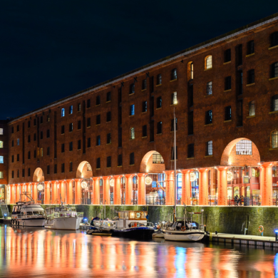 The Tate Liverpool building at the Royal Albert Dock illuminated at night. The historic red-brick warehouse is lit with warm orange lighting along its colonnade, while boats are moored in the dock in the foreground. Reflections of the building’s lights shimmer across the water, creating a colourful waterfront scene against a dark evening sky.