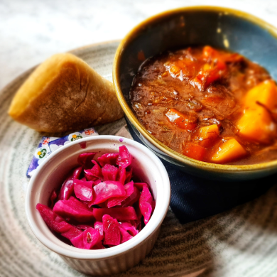 A plated meal featuring a bowl of hearty beef stew with chunks of potato and vegetables, served alongside a bread roll and a small dish of pickled red cabbage. The food is presented on a large ceramic plate on a marble tabletop, with warm lighting highlighting the rich colours of the dish.