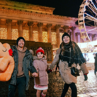 Family walking through Liverpool Christmas Market at night, holding a large festive plush toy, with St George’s Hall illuminated in the background and a brightly lit Ferris wheel towering above the market stalls. Wet cobbled streets reflect the colourful lights of the festive scene.