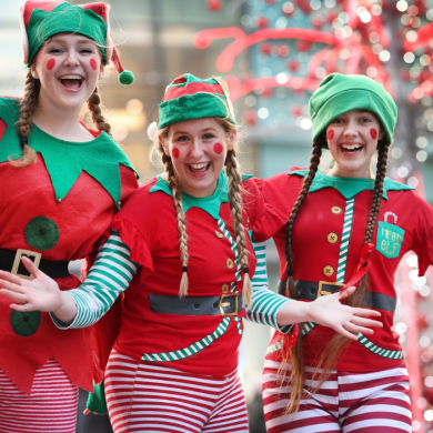 Three performers dressed in festive elf costumes pose in a city centre setting, wearing red and green outfits with striped sleeves and tights. Decorative Christmas lights and illuminated seasonal displays create a colourful blurred backdrop behind the group.