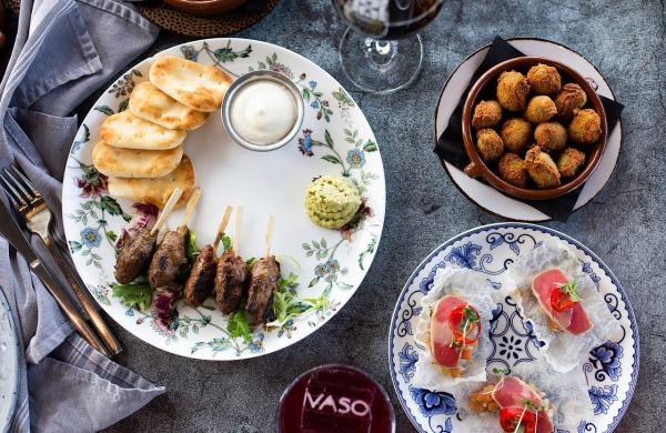 A table setting with grilled kebabs, pita bread, sauce, fried bites, and garnished appetizers served on floral-patterned plates at VASO.
