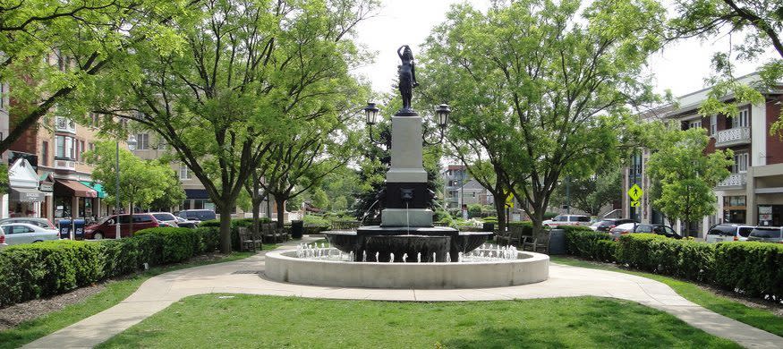 Image is of the Hyde Park statue that has water shooting out into a pool in the center of a town that splits the road with buildings on each side.