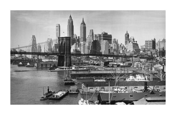 Black-and-white mid-20th-century photograph of the Brooklyn Bridge and Lower Manhattan skyline as seen from the Brooklyn waterfront, with commercial ships, barges, and dockside warehouses lining the East River in the foreground.