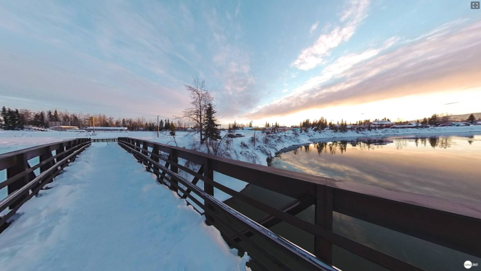 a panoramic view from a snow-covered pedestrian bridge over a river with a sunset colored sky
