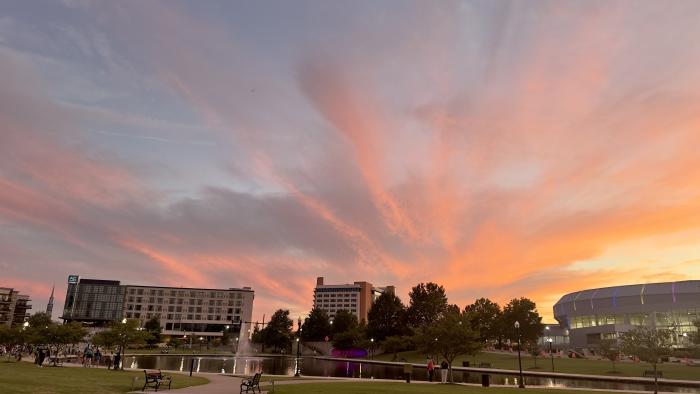 A vibrant sunset fills the sky with shades of pink, orange, and purple over Big Spring Park in downtown Huntsville. The park's pond, fountain, walking paths, and surrounding skyline create a colorful evening scene as visitors enjoy the park.