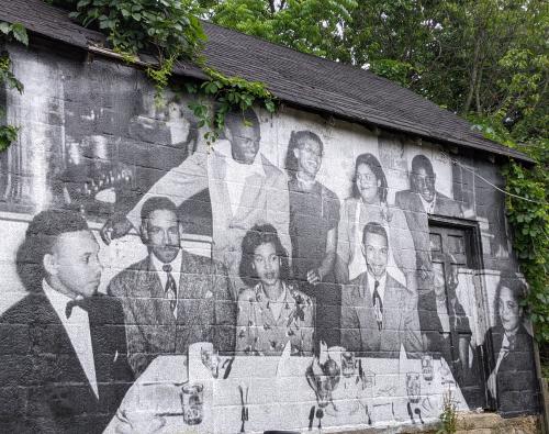 A small building has a mural covering the entire wall depicting a black and white photo of a group gathered around a table smiling.