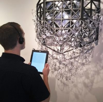 A young Caucasian man with short brown hair wearing headphones holds an iPad up to a metallic silver geometric sculpture to listen to an audio description.  The artwork pictured is by former Universal Access Artist in Residence, Katie Shaw