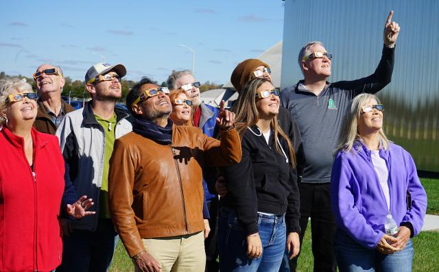 A group of individuals, wearing solar eclipse glasses and looking to the sky, as if they are viewing a solar elcipse.