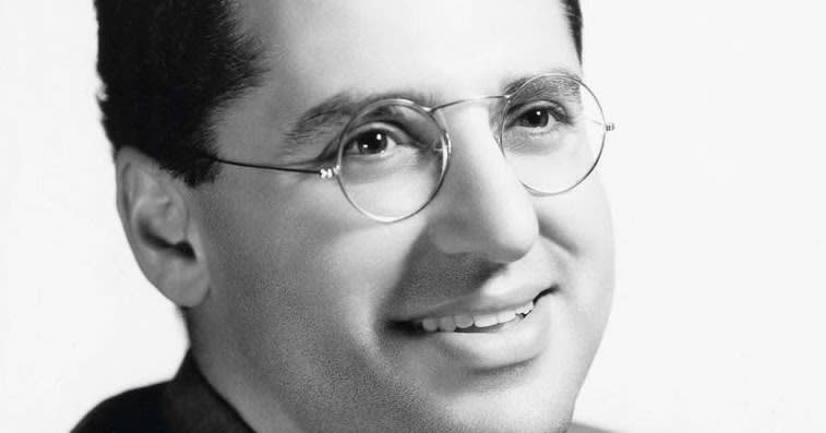 Black and white portrait of a smiling man wearing round glasses and a suit jacket.