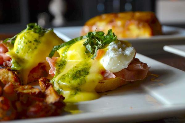 Close-up of a gourmet eggs Benedict dish served on a white plate, featuring poached eggs topped with hollandaise and green herb sauce, garnished with fresh basil and set atop prosciutto and toasted bread. Crispy breakfast potatoes and another brunch item are visible in the background.