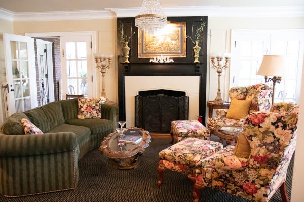 An image of a cozy living room featuring two chairs with floral upholstery and a green couch. Fireplace in the center of the image with French doors on either side. Lighting is bright from a sun coming through windows and glass.