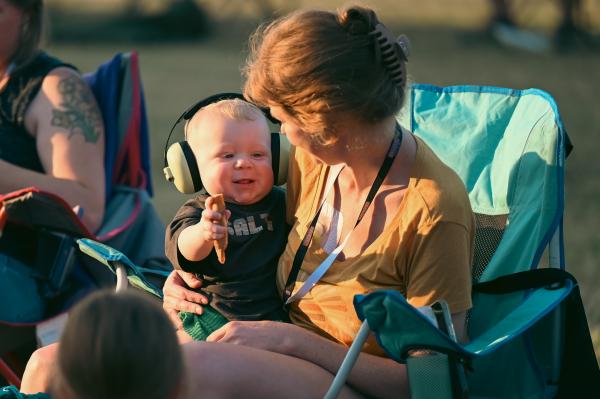 A baby sits in his mothers lap while holding onto an ice cream cone and wearing ear muffs