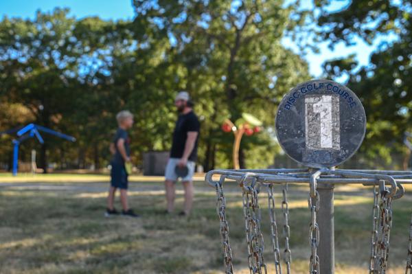 A man and child prepare to throw a frisbee into the first disc golf basket on a sunny afternoon at Ben Geren Park in Fort Smith, Arkansas.