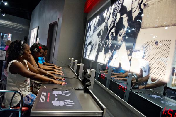 Black women sitting at a 1950s lunch counter inside a museum with headphones on while watching historical film