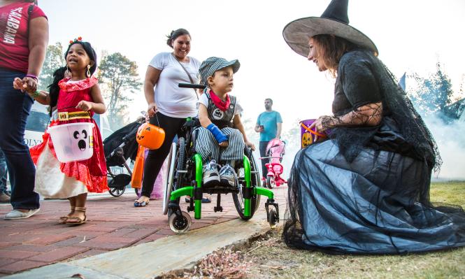 Adorable child in wheelchair with his guardian and Spooky Springs Witch character trick-or-treating.