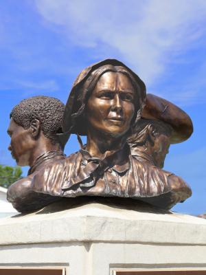 A bronze statue depicts three pioneers facing away from each other, the woman pioneer faces the camera, wearing colonial garb.