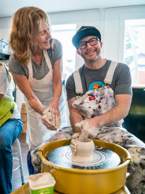 Man at a pottery wheel spinning clay while woman stands smiling beside him