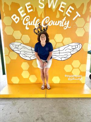 a girl poses in front of a yellow mural with wings and words "bee sweet Gulf County"