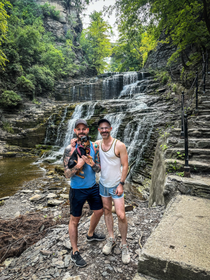 Two men stand posing with a small dog being held in front of a waterfall.