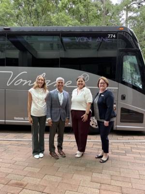 Four people smile at the camera, standing in front of a luxury bus. From left to right: (a blonde woman in a white blouse and gray slacks), Alex Danza (Founder and CEO of Vonlane, a man wearing a gray plaid suit), Elizabeth Eddins (Executive Director of Visit The Woodlands, a woman wearing a white blouse and maroon slacks), and Jennifer Gohagan (GM of The Woodlands Waterway Marriott, wearing a patterned shirt with a navy sweater and slacks). The bus behind them is long, silver and black, and has "Vonlane" written in smooth cursive across the side.