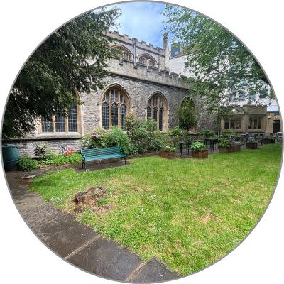 An circular shaped image showing St Stephens Church Garden Quiet Area. There is a grassy area with benches and flower pots alongside in most of the photo. At the back there is a Church with the stained glass window shape showing from the outside.