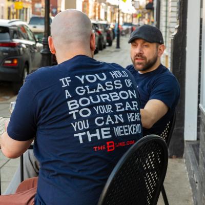 A man sits facing away from the camera. The back of his shirt reads "If you hold a glass of bourbon to your ear you can hear the weekend". Behind him is another man, facing the camera and looking at the first man.