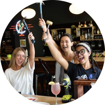 Three women holding up hammers and mallets with cocktails on the table