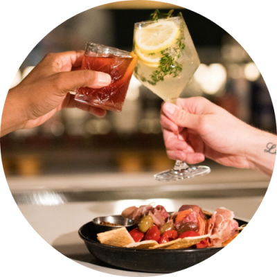 Two alcoholic drinks clinking over tray of appetizers