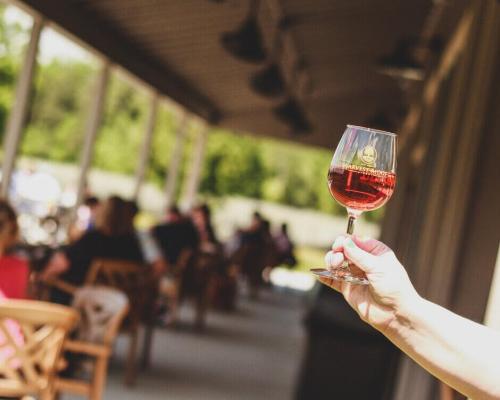 A hand holding a glass of wine with a blurred Harvest Ridge Winery in the background.