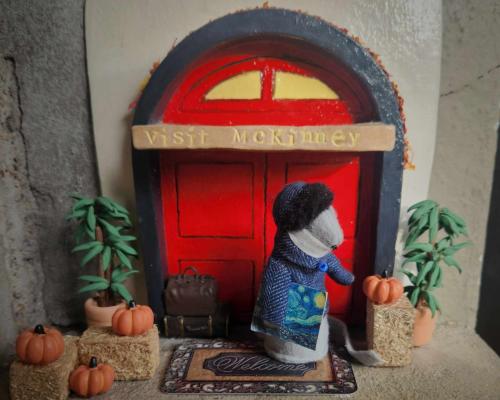 Tiny door in red with pumpkins and hay bales with a small cloth mouse standing in front of it holding a painting