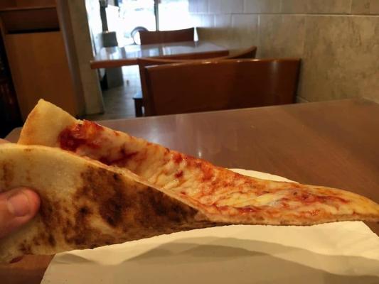 A hand holding thin, large triangular New York-style cheese pizza slice together, showing the charred underside crust and melted mozzarella topping, inside a pizza shop.
