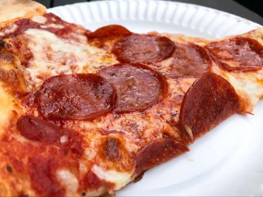 Close-up of a large New York-style pepperoni pizza slice on a white paper plate, loaded with cupped, crisped pepperoni rounds over melted mozzarella and tomato sauce.