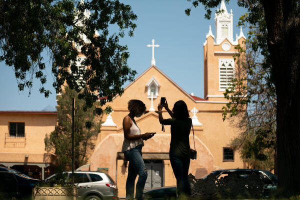 Two people holding phones stand in front of the San Felipe de Neri Church in Old Town Albuquerque