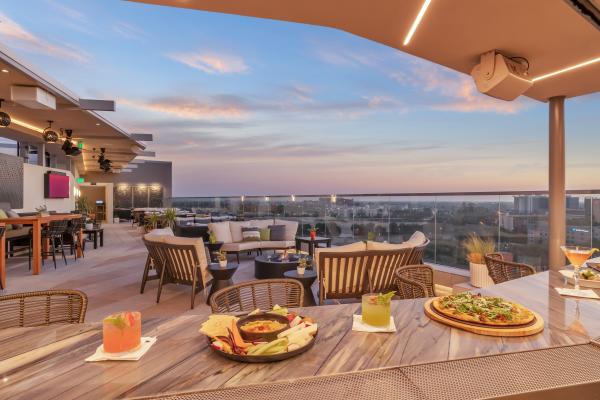 Image of a charcuterie board, a pizza topped with greens and two cocktails placed on top of the bar at Parkestry. In the background are lounge seating options and a view of the Anaheim skyline.