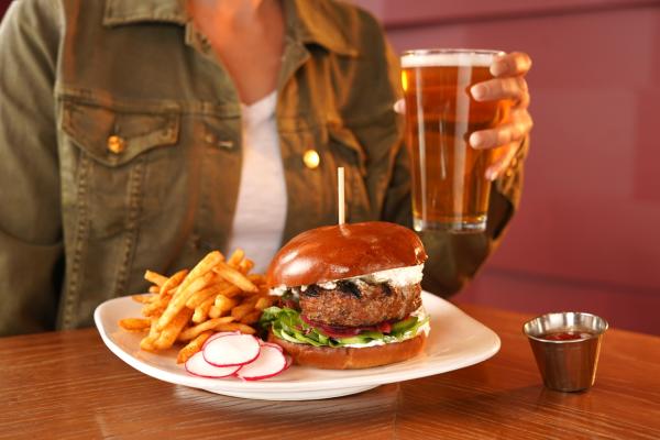 Image of a burger on a white plate, served with a side of fries. In the background, a woman in a green jacket and white shirt is hold a beer, next to the burger pictured in the foreground.