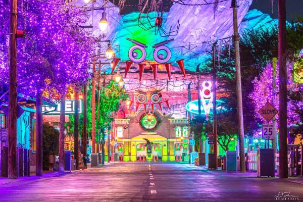 Image of Cars Land inside Disney California Adventure Park decorated for Halloween. Image shows the main walkway of Cars Land with tires and traffic cones hung in the air, above the walkway. They are designed to look like scary faces. Image is taken at night and includes light up signs and decor that are red, purple, yellow, green and orange.