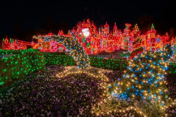 Image of it's a small world at Disneyland, all lit up with holiday lights at night. Red lights can be seen twinkling from the attraction's facade. In the foreground, bushes can also be seen with holiday lights.
