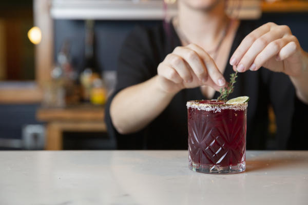 A deep purple cocktail with a salt rim sits on a bar. A bartender puts herbs in it.