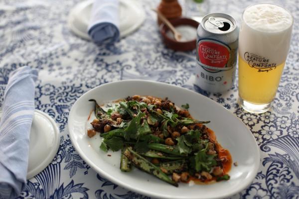 A plate of okra and chickpeas with seasonings and accent flavors on a white plate sits on a blue and white table cloth. A can and glass of Creature Comforts beer sits beside it.