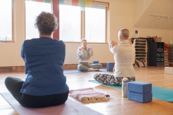 Three individuals practice yoga in a serene studio, with a focus on a seated instructor guiding the session. Natural light filters through large windows, enhancing the calming atmosphere.