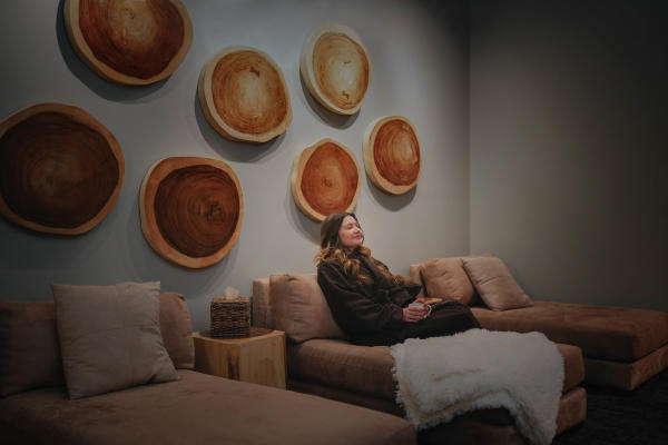A serene lounge area features a woman relaxing on a plush sofa, surrounded by wooden wall art and cozy decor elements.