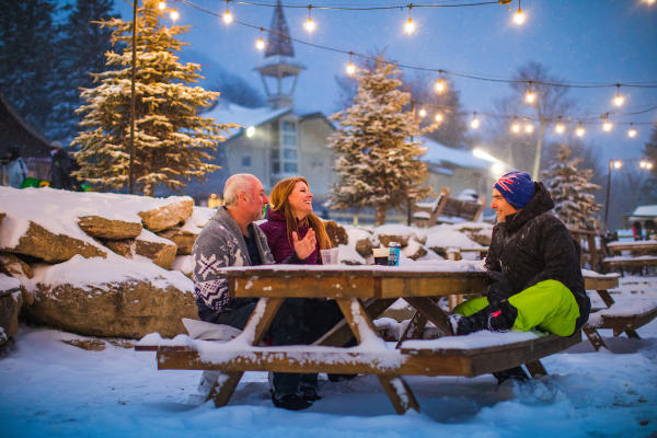 A group of three people dressed warmly sit at a picnic table with a light snow cover. Snow covered trees and string lights are behind the group.